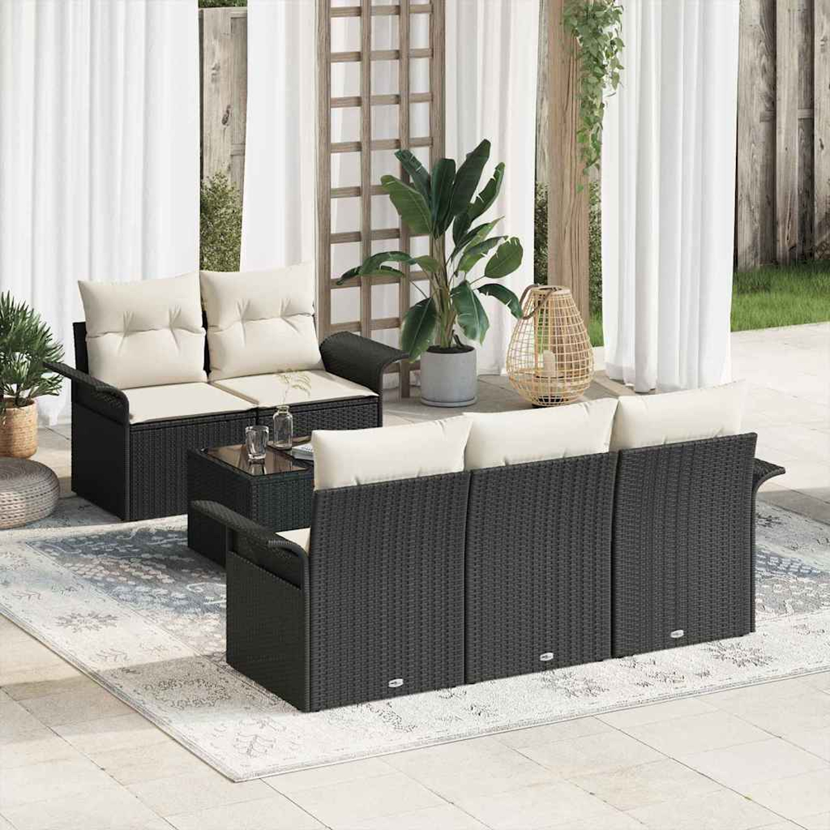 Terrassen-Set im Freien mit schwarzer Korbmöbeln, cremefarbenen Kissen, Glastisch, Teppich, Pflanzen und weißen Vorhängen. Hintergrund ist ein Garten.