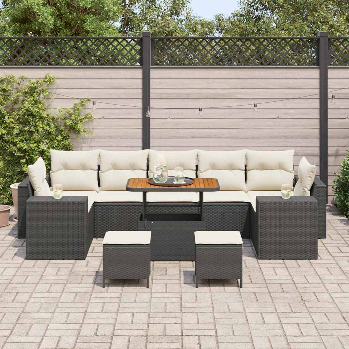 Juego de patio al aire libre con muebles de mimbre gris oscuro, cojines beige y una mesa pequeña. Una valla y vegetación en el fondo.