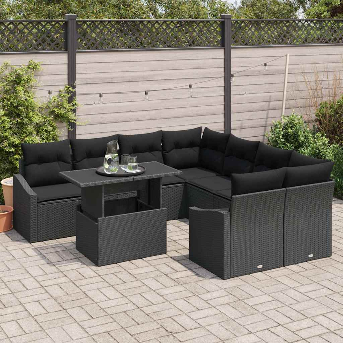 Eine Terrassengarnitur mit schwarzen Kissen, einem Tisch mit Getränken und einem Zaun im Hintergrund. Das Set steht auf einer gefliesten Terrasse.