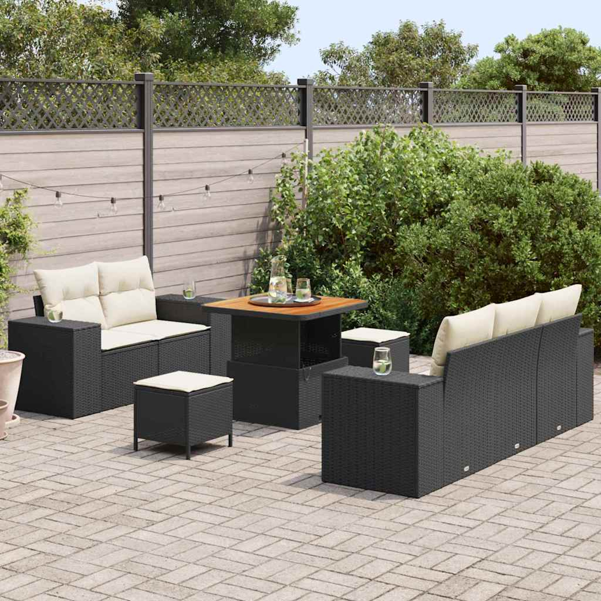 Garten-Terrassenset: Schwarze Korbmöbel mit cremefarbenen Kissen, quadratischer Tisch und Hocker auf gepflasterter Terrasse. Grüne Büsche und Zaun.
