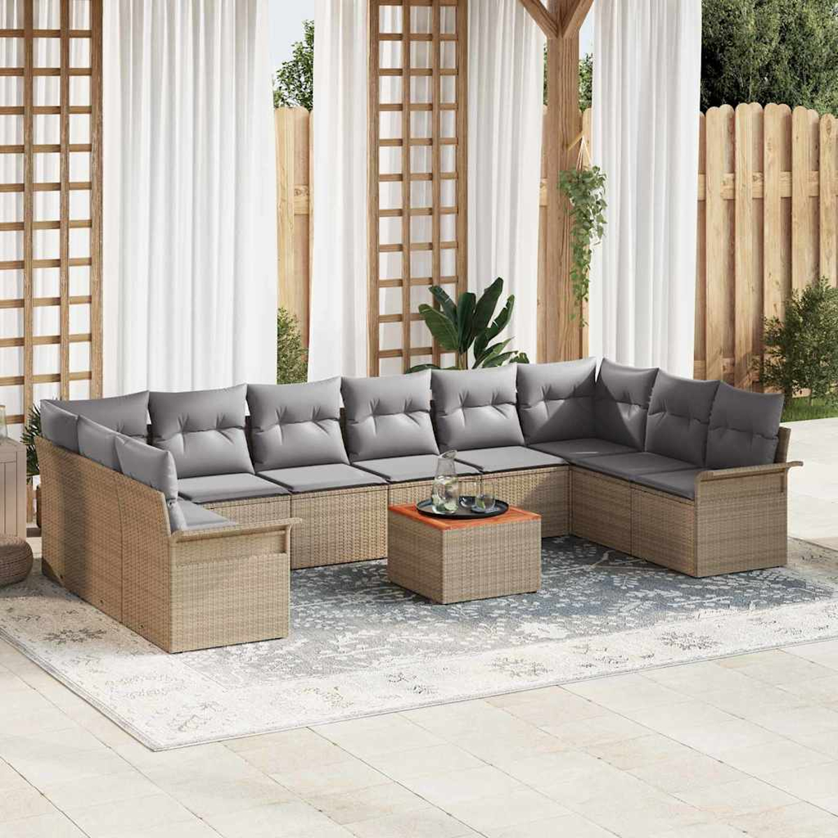 Outdoor-Ecksofa mit grauen Kissen auf einem gemusterten Teppich. Ein kleiner Tisch mit Getränken steht davor. Weiße Vorhänge und ein Holzzaun sind dahinter.