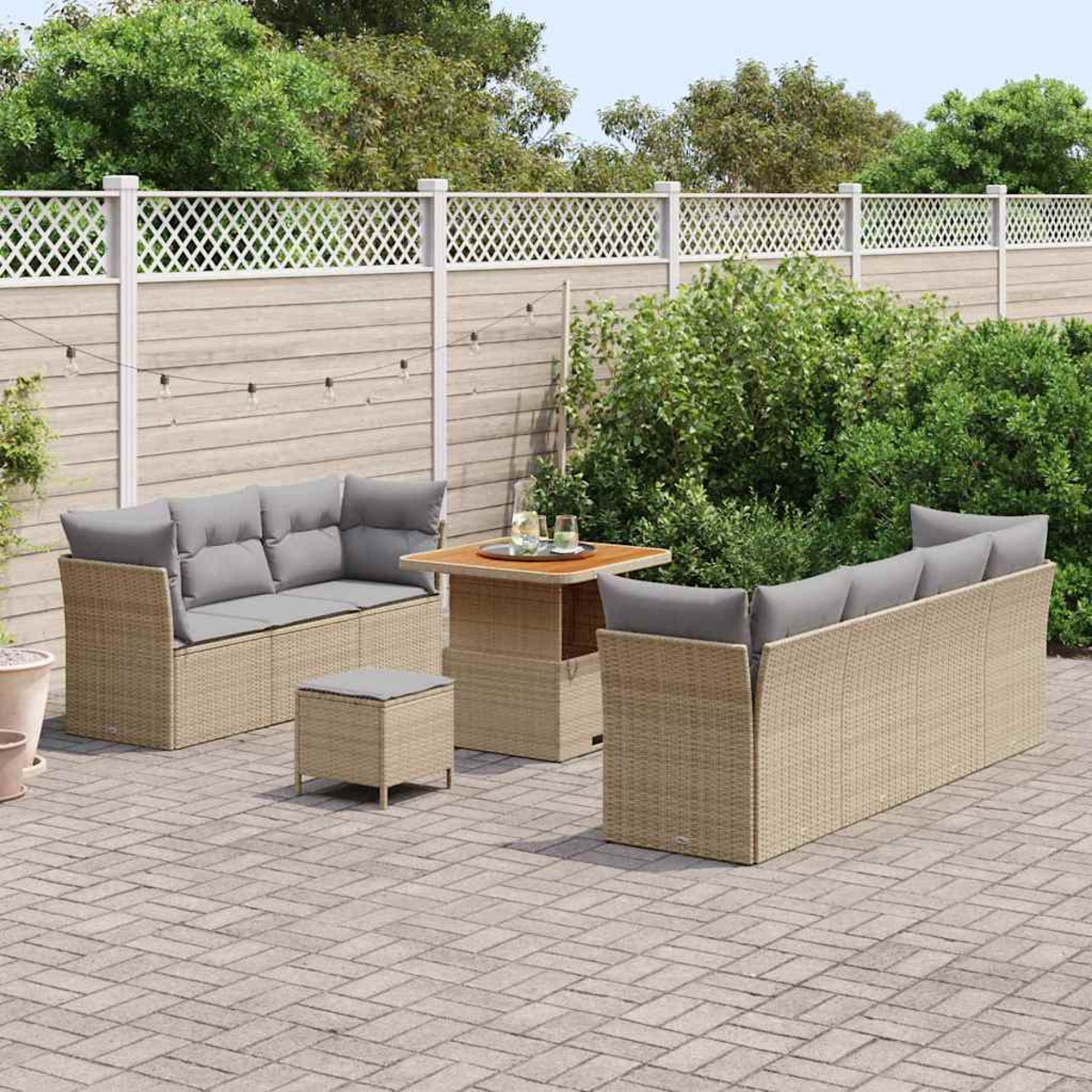 Set divano in rattan da esterno con cuscini grigi, tavolo e pouf su un patio in mattoni. Una recinzione bianca e vegetazione sullo sfondo.