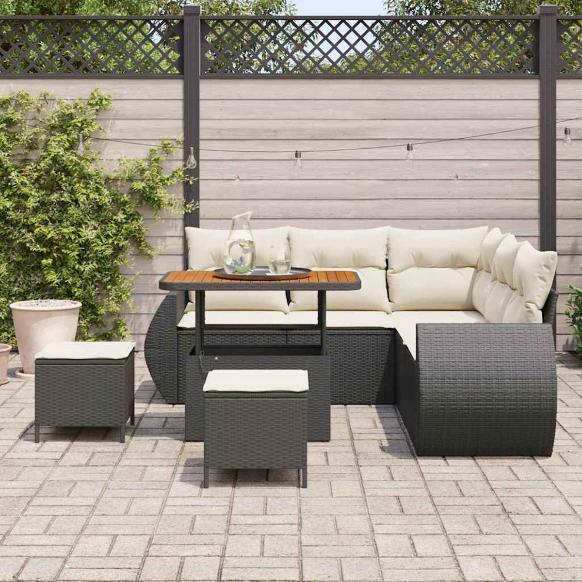 Terrassenmöbel-Set mit Ecksofa, kleinem Tisch und zwei Hockern. Auf dem Tisch stehen ein Krug und Gläser. Im Hintergrund ist ein Zaun.