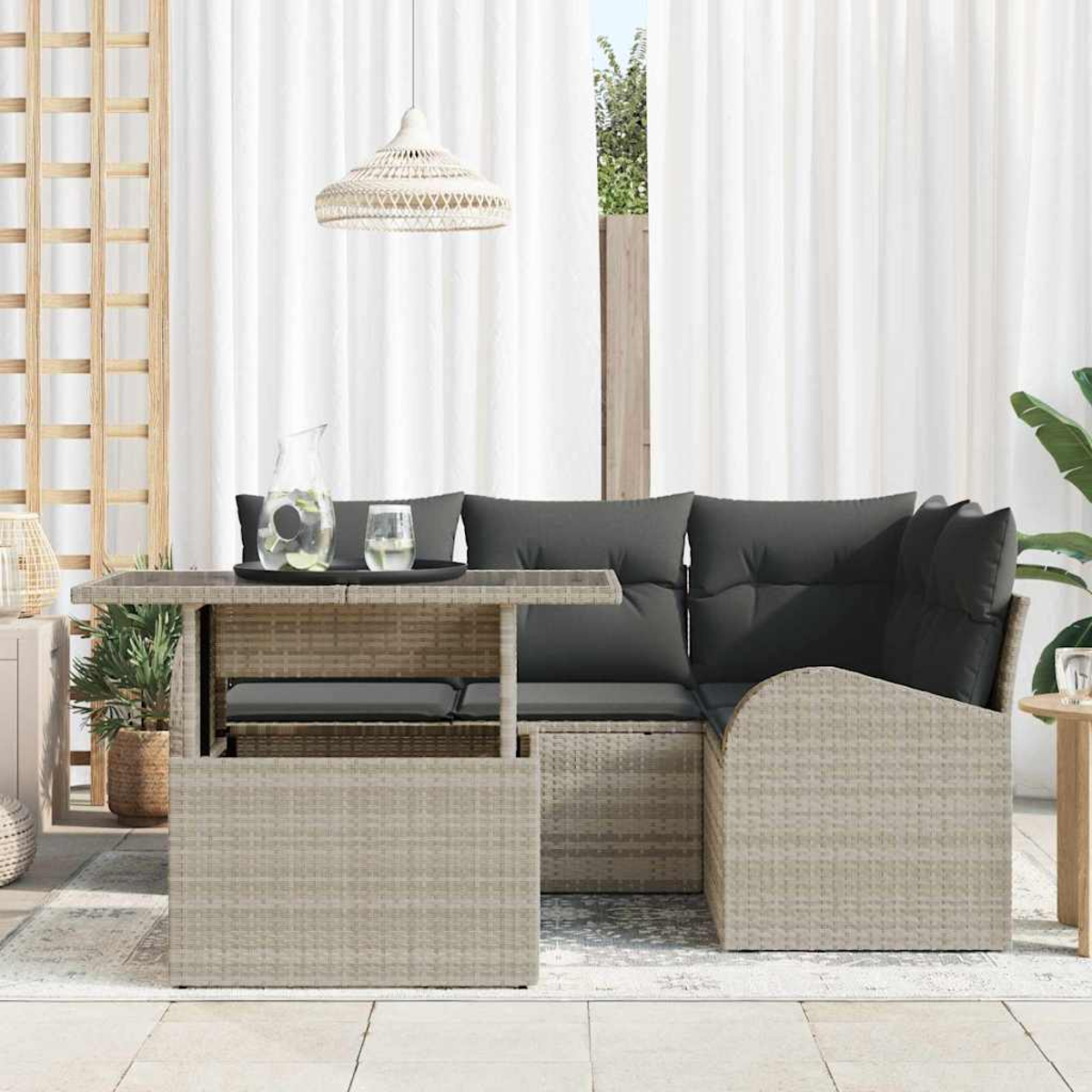 Szene im Freien mit einem Korbtisch und Ecksofa mit grauen Kissen. Auf dem Tisch stehen ein Krug und Gläser. Weiße Vorhänge im Hintergrund.