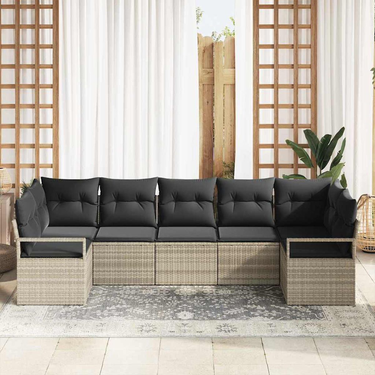Ein beiges Ecksofa mit schwarzen Kissen steht auf einer Terrasse. Darunter liegt ein grauer Teppich. Weiße Vorhänge und Holzgitter sind im Hintergrund.