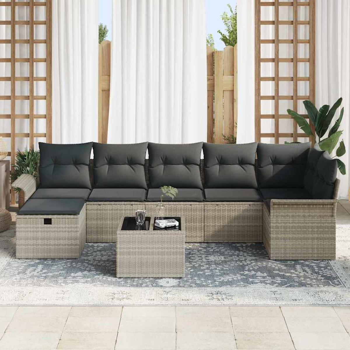 Außenbereich. Graue Kissen auf hellbraunem Rattan-Sofa, kleiner Tisch, Teppich, weiße Vorhänge und Pflanzen.