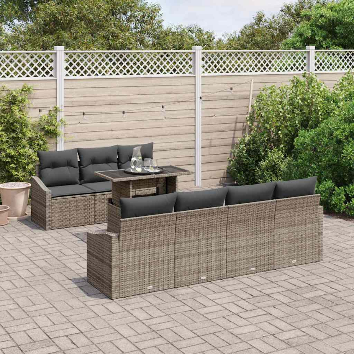 Geflochtenes Gartenmöbel-Set auf einer Terrasse. Beinhaltet ein Sofa, eine Doppelsitzcouch und einen Tisch. Ein weißer Zaun und grünes Laubwerk im Hintergrund.