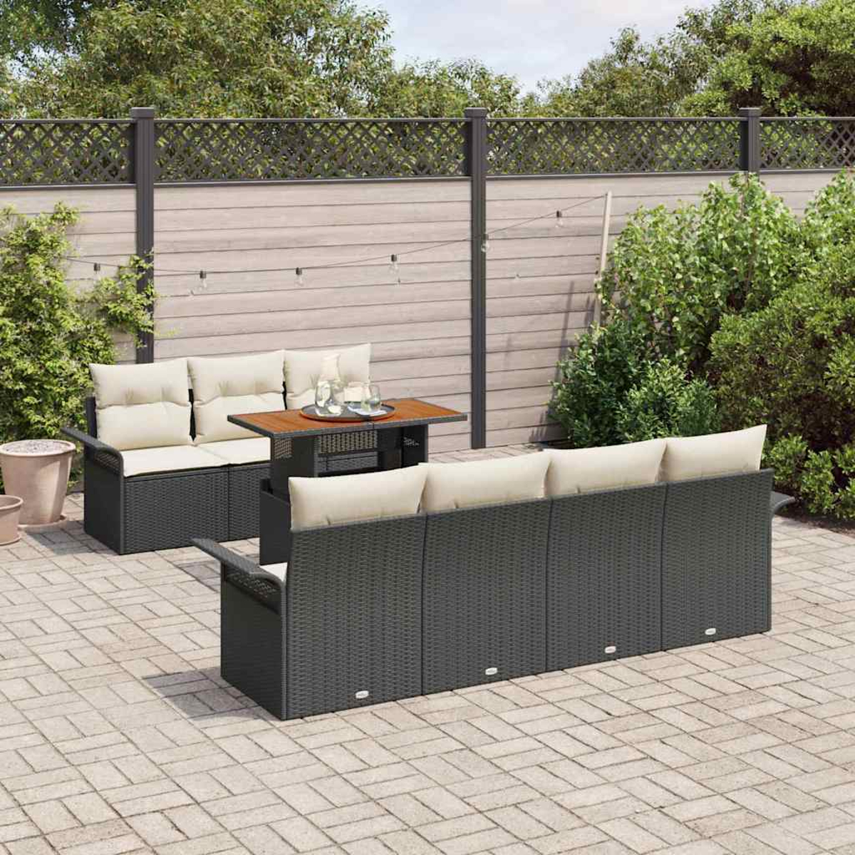 Outdoor-Terrassenset mit schwarzen Korbmöbeln, beigen Kissen und einem Holztisch. Ein Zaun und grüne Pflanzen befinden sich im Hintergrund.