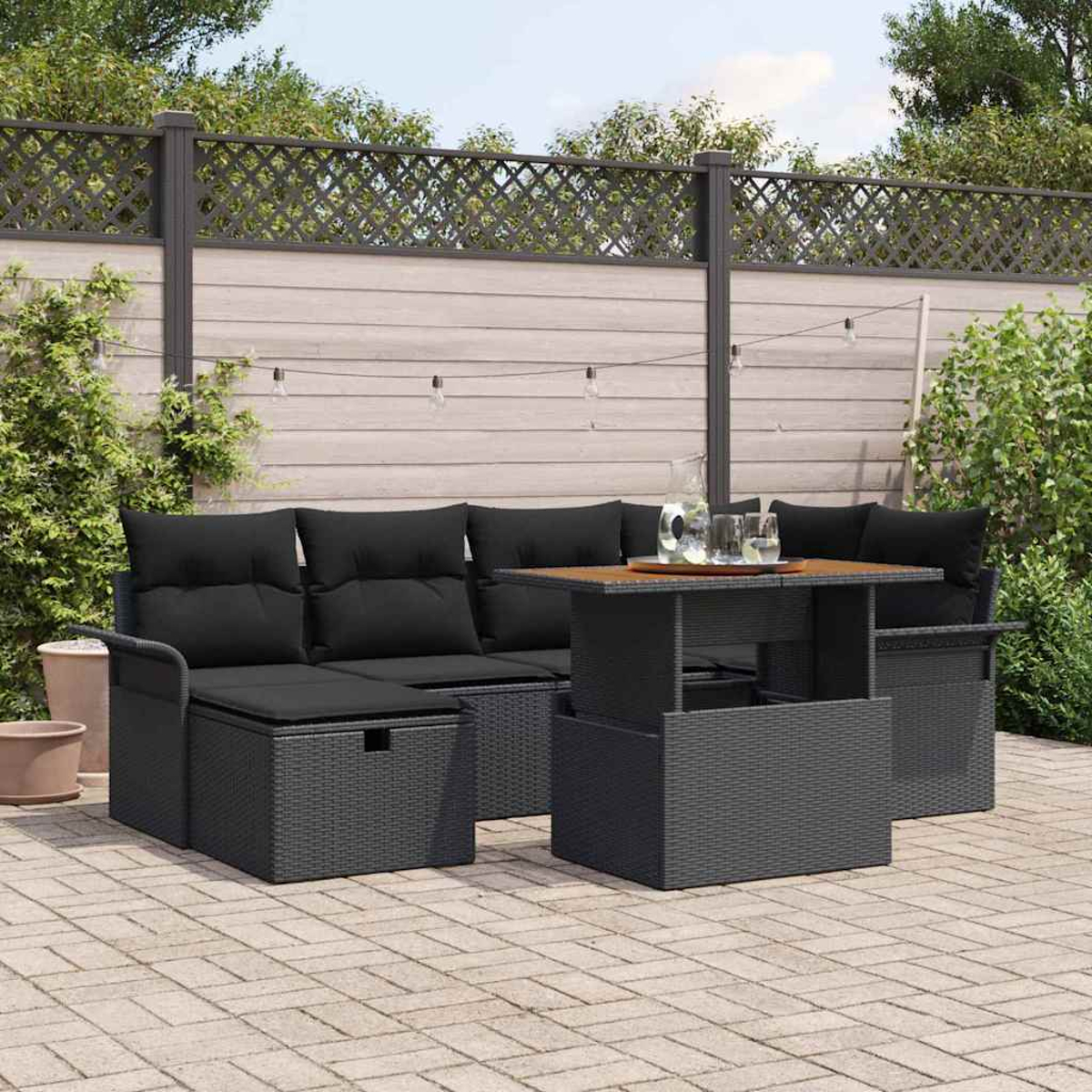 Garten-Sitzgruppe. Schwarzes Korbmöbel-Sofa und Tisch mit Hubfunktion auf einer Terrasse. Hintergrund: Zaun, Pflanzen, Lichterkette.