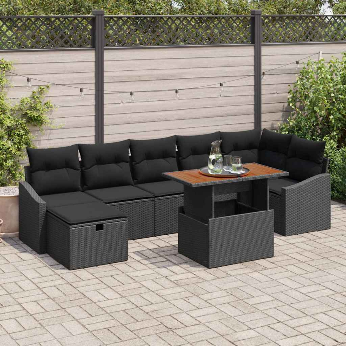 Garten-Terrassenset: schwarzes Korbmöbel-Sofa mit Kissen, Tisch, Wasserkrug, Gläser. Auf Steinterrasse, Holzzaun-Hintergrund.