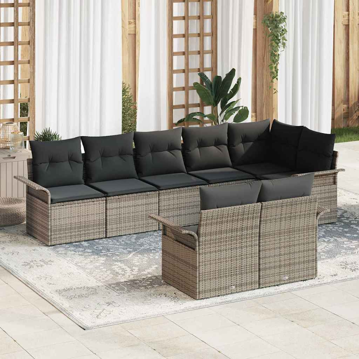 Geflochtenes Outdoor-Ecksofa mit dunklen Kissen auf einem gemusterten Teppich. Holzzaun und weiße Vorhänge im Hintergrund mit Pflanzen.