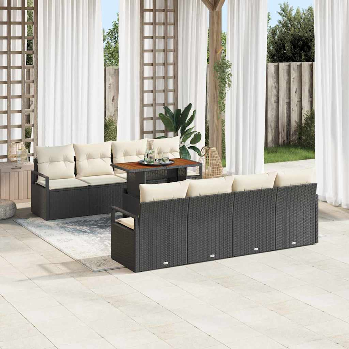 Juego de muebles de patio al aire libre con sofás de mimbre negro, cojines crema y una mesa de madera. Cortinas blancas y vegetación al fondo.