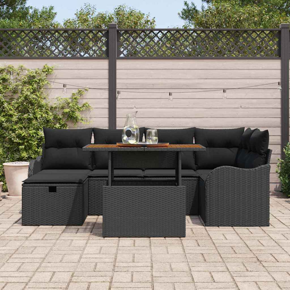 Schwarzes Outdoor-Wickermöbelset mit Kissen und einem Tisch. Im Hintergrund sind ein Zaun, Pflanzen und Lichterketten.