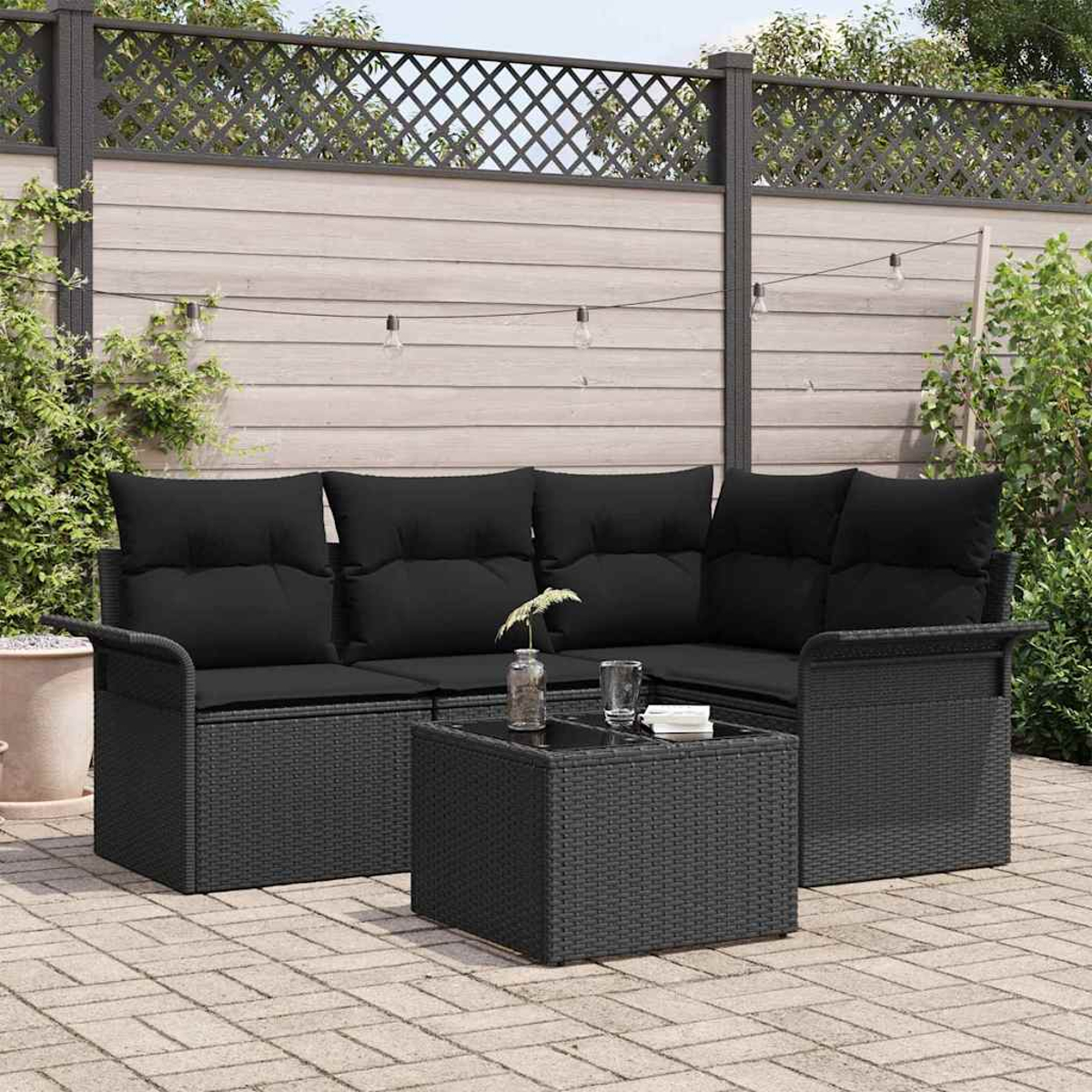 Schwarzes Korbmöbel-Set für die Terrasse. Es umfasst ein Ecksofa und einen Couchtisch. Der Hintergrund zeigt einen Holzzaun und Grün.
