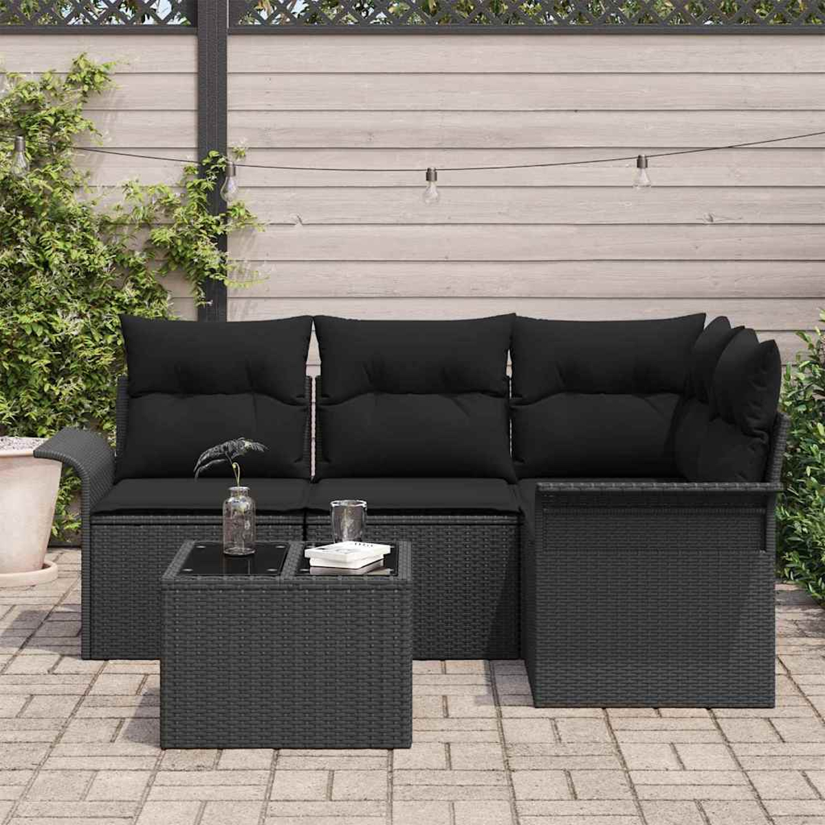 Ein schwarzes Korbmöbel mit schwarzen Kissen und einem kleinen Tisch mit Glas und Buch steht auf einer Ziegelterrasse.