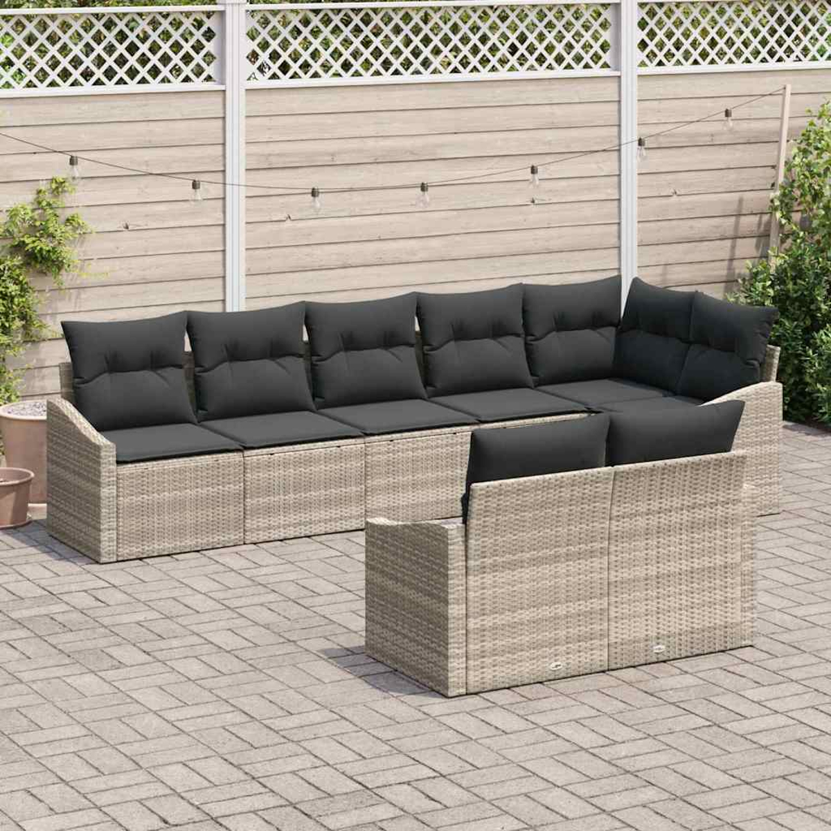 Ein Outdoor-Korbmöbel mit schwarzen Kissen steht auf einer Ziegelterrasse. Im Hintergrund ein weißer Zaun und eine Holzwand.