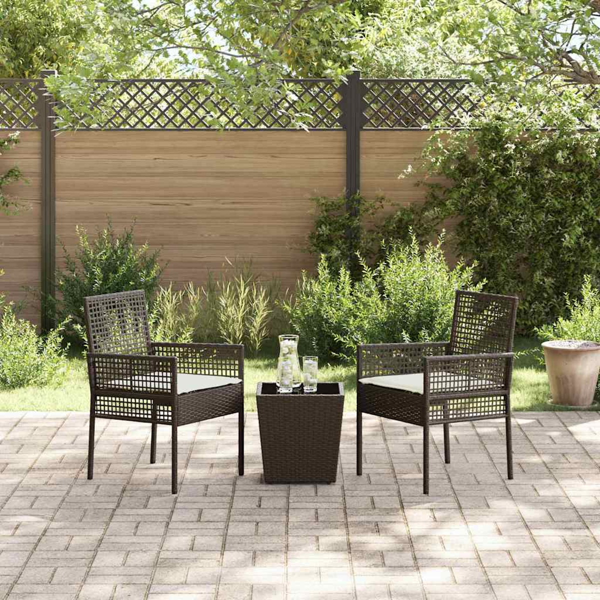 Juego de patio exterior con dos sillas, una mesa pequeña y un vaso de agua. Arbustos verdes y una valla de madera al fondo.