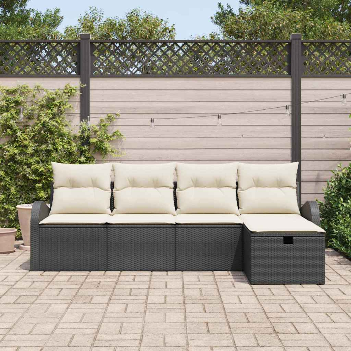 Ein graues Outdoor-Ecksofa mit cremefarbenen Kissen steht auf einer Backstein-Terrasse. Im Hintergrund ist ein Holzzaun zu sehen.
