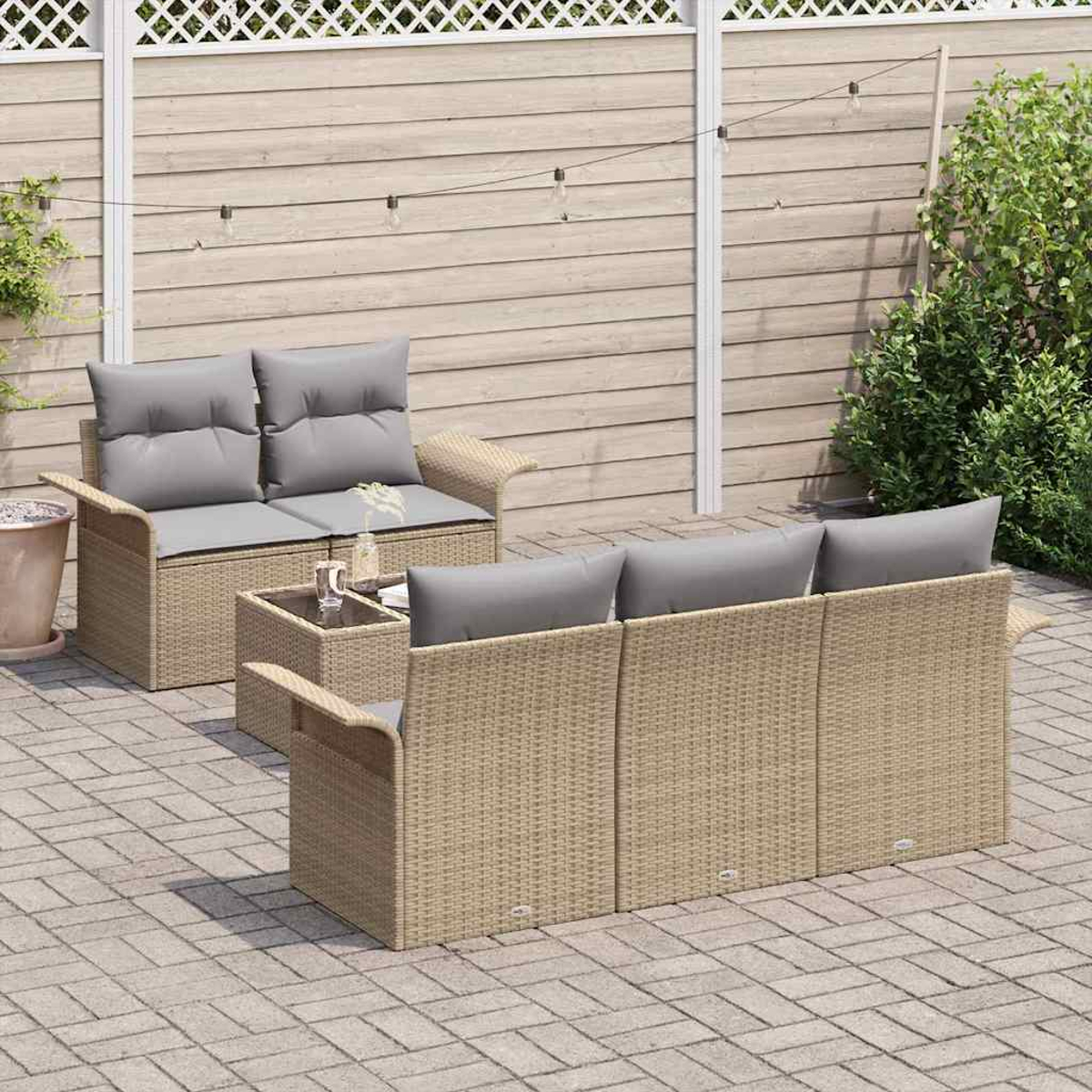 Gartenmöbelset aus Korbgeflecht mit grauen Kissen, Couchtisch, auf einer Backstein-Terrasse, vor Holzzaun und Grün.