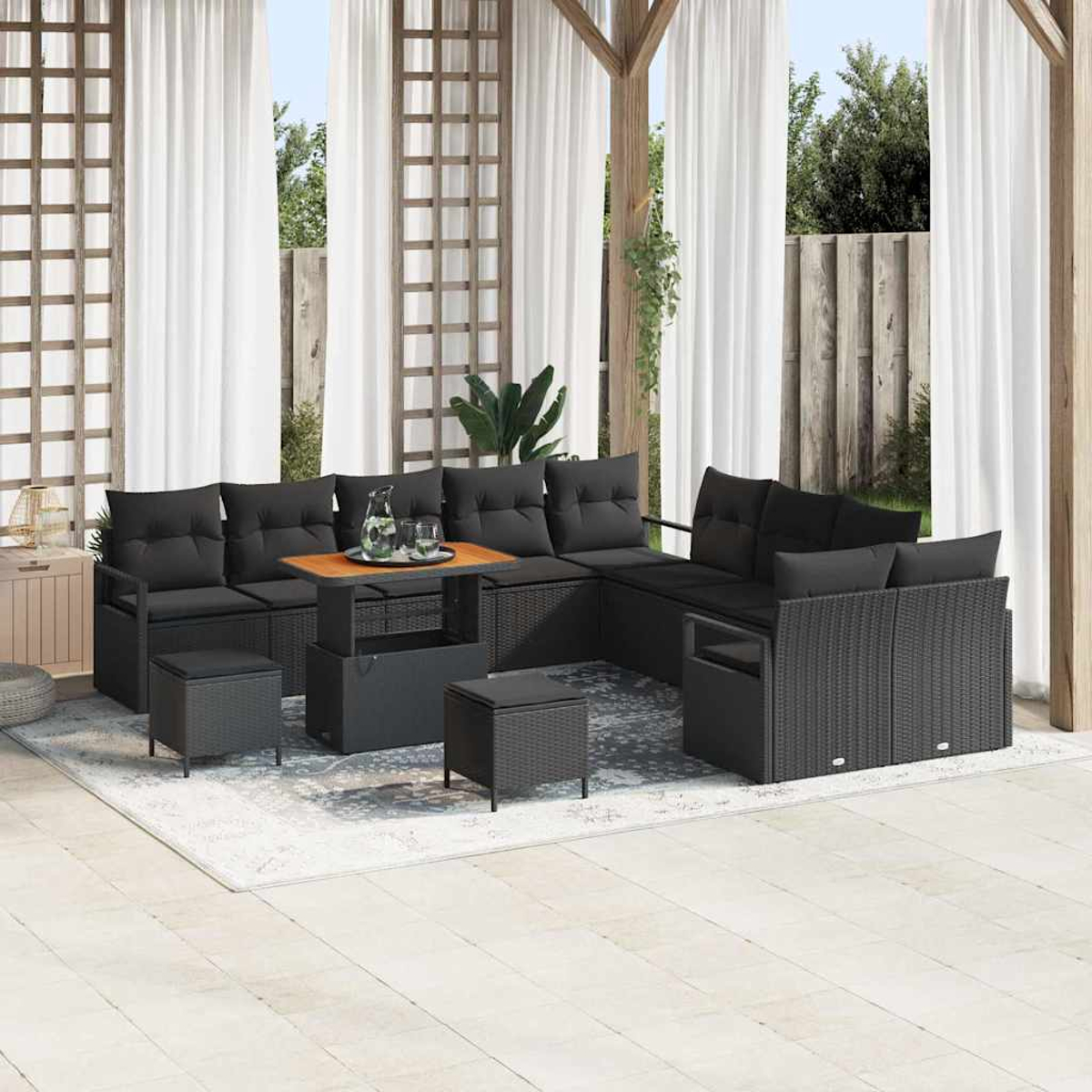 Schwarzes Outdoor-Wicker-Ecksofa mit Kissen, einem Holztisch und Ottomanen auf einem Teppich unter einer Pergola mit weißen Vorhängen.