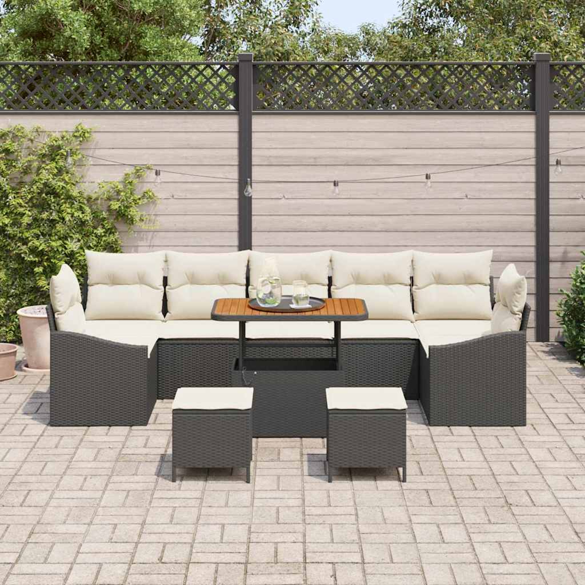 Garten-Sitzgruppe mit Tisch, Kissen und zwei Hockern. Das Set steht auf einer Ziegelterrasse, im Hintergrund ein Zaun und Grün.
