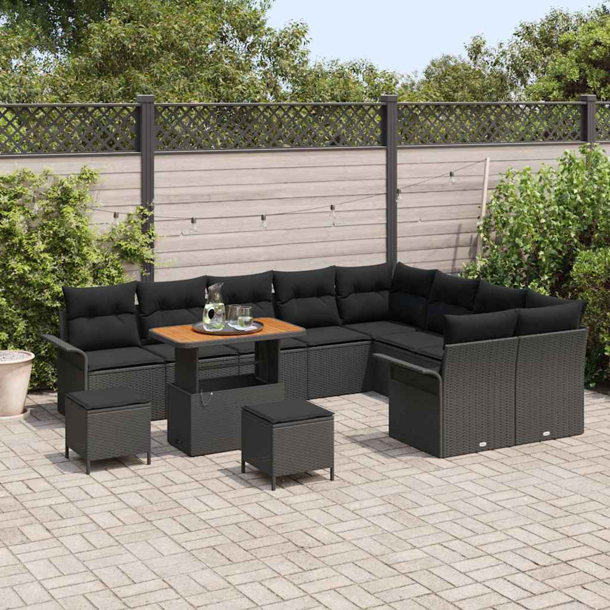 Outdoor-Ecksofa mit schwarzen Kissen, kleinem Tisch und Hockern. Auf einer Terrasse mit Holzzaun und Grün im Hintergrund.