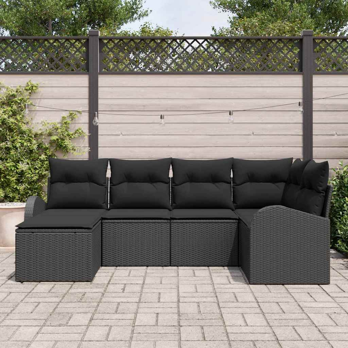 Ein dunkelgraues Outdoor-Ecksofa aus Korbgeflecht mit schwarzen Kissen steht auf einer Ziegelsteinterrasse. Ein Zaun und Grün bilden den Hintergrund.