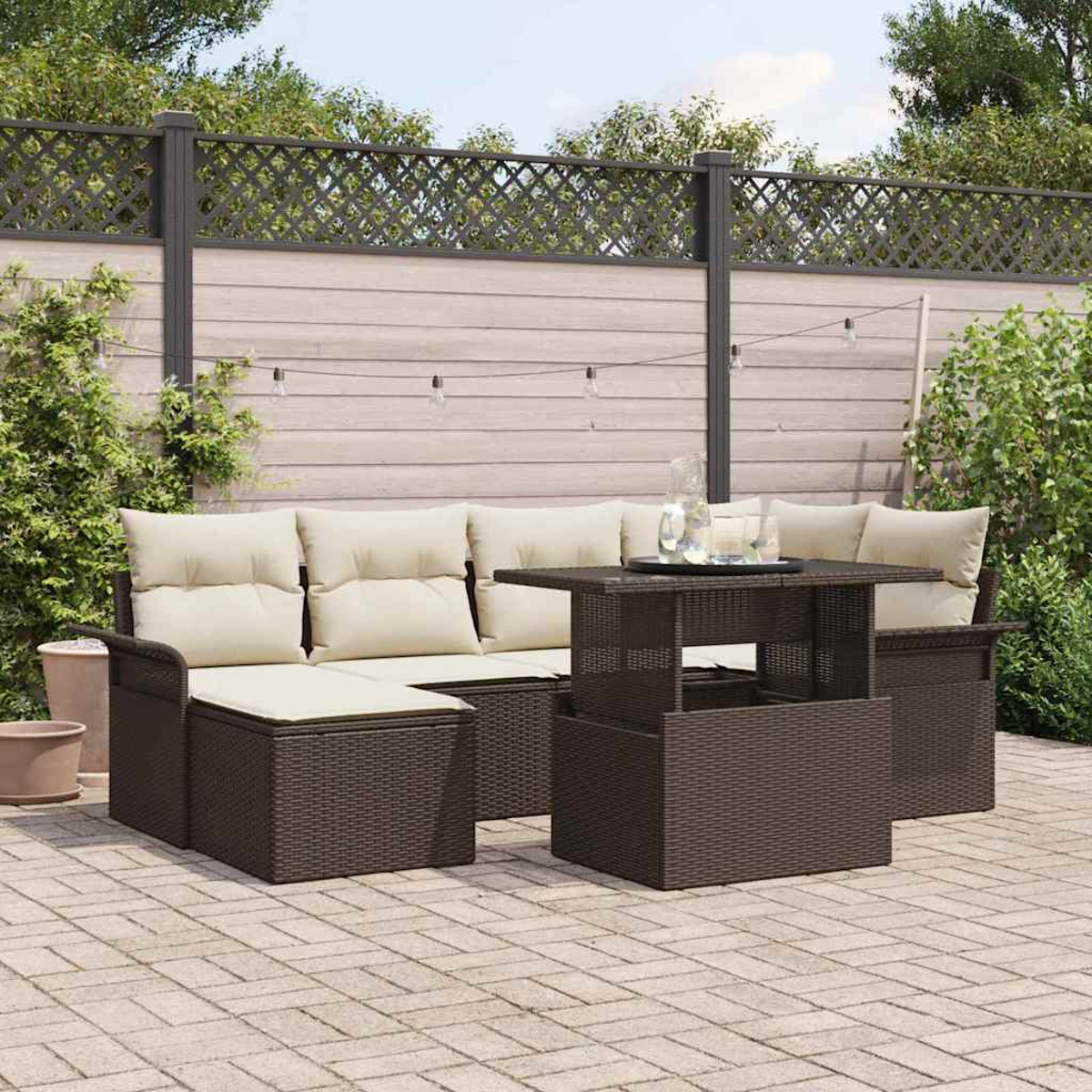 Terrassenmöbel: braunes Korbssofa mit cremefarbenen Kissen, Tisch und Ziegelpflaster. Hintergrund: Zaun und Grün.