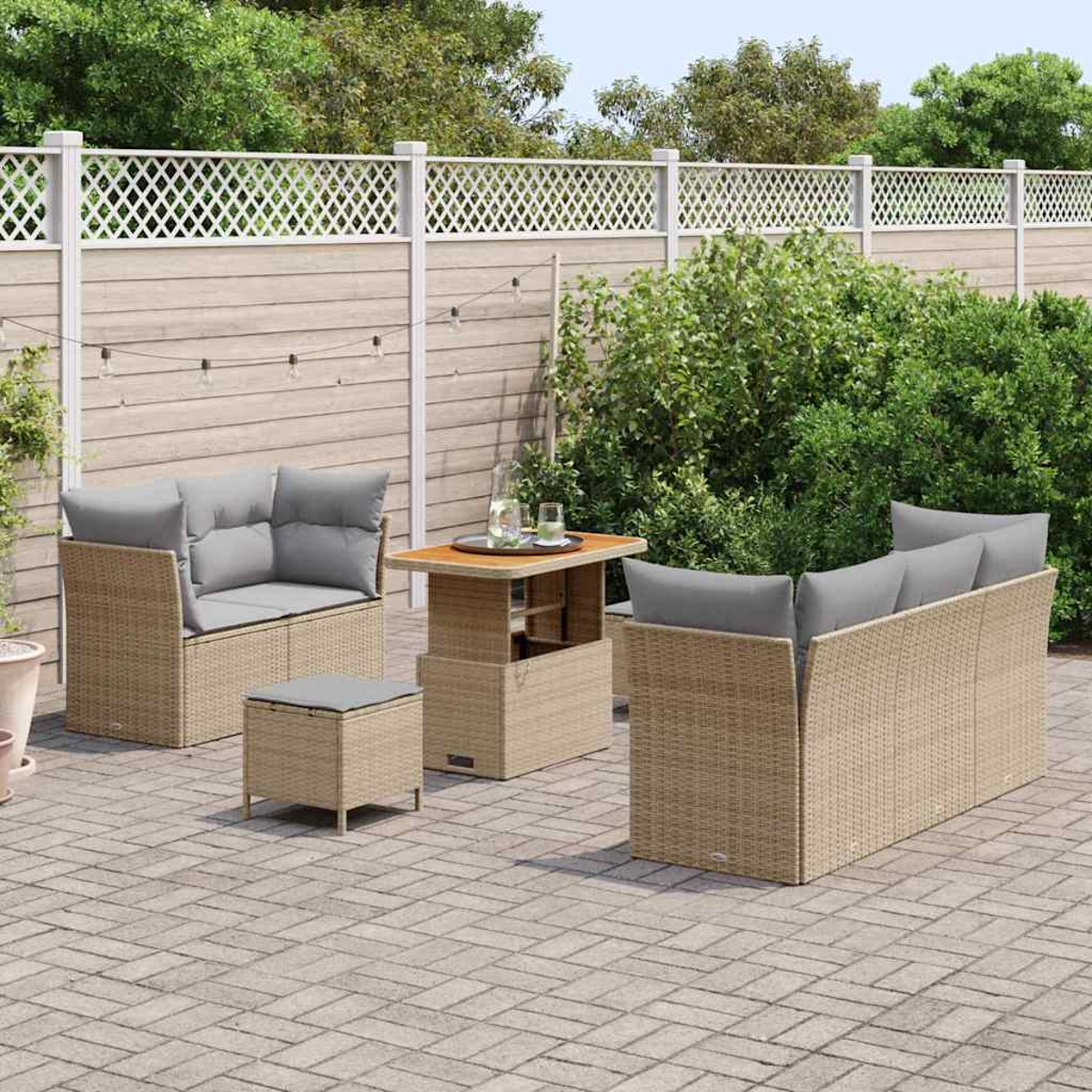 Set da patio esterno con mobili in vimini beige, cuscini grigi e tavolo in legno. Recinzione a traliccio bianca e verde lussureggiante.