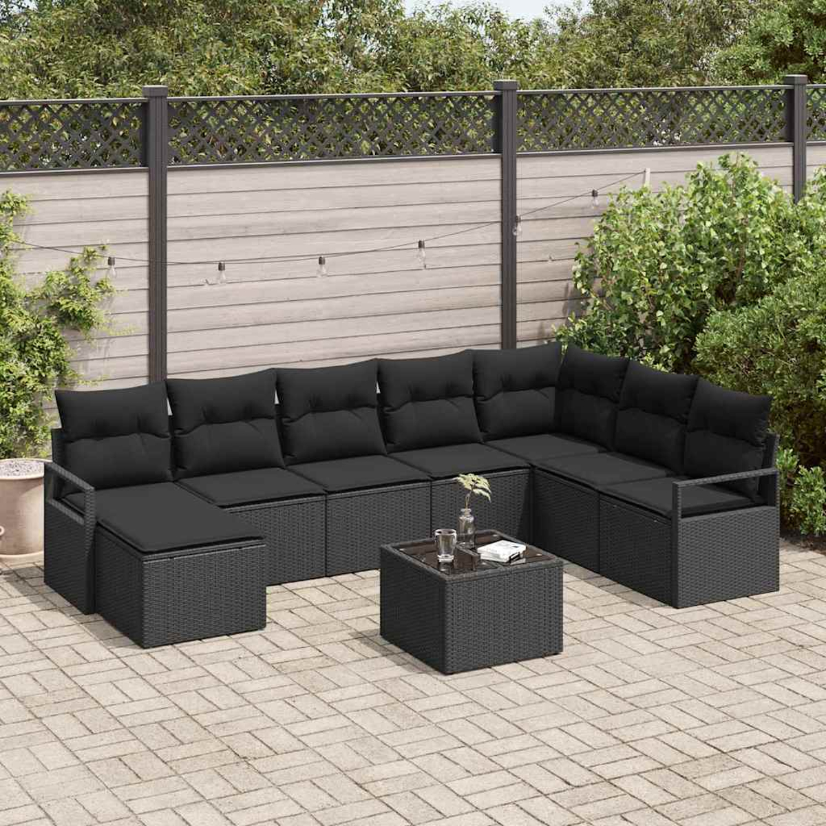 Schwarzes Outdoor-Ecksofa aus Korbgeflecht mit Kissen, kleiner Tisch mit Glas und Backstein-Terrasse. Grüne Pflanzen und Holzzaun im Hintergrund.