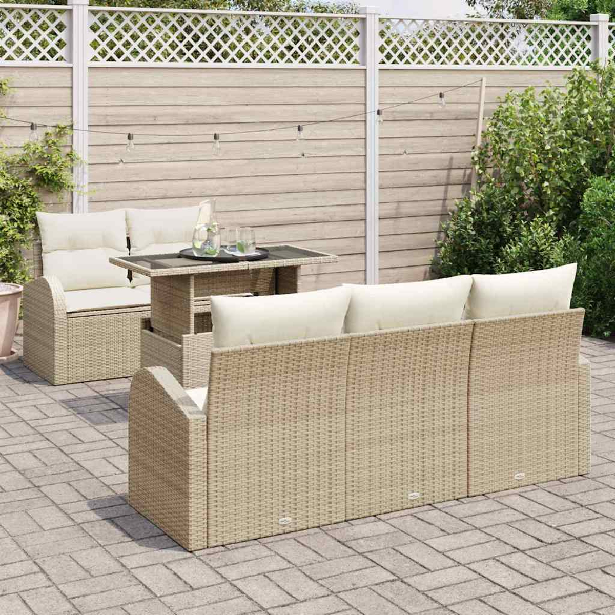 Terrassenset mit beigen Korbmöbeln, weißen Kissen und einem Tisch mit Glasplatte. Ein weißer Zaun und Grün im Hintergrund.