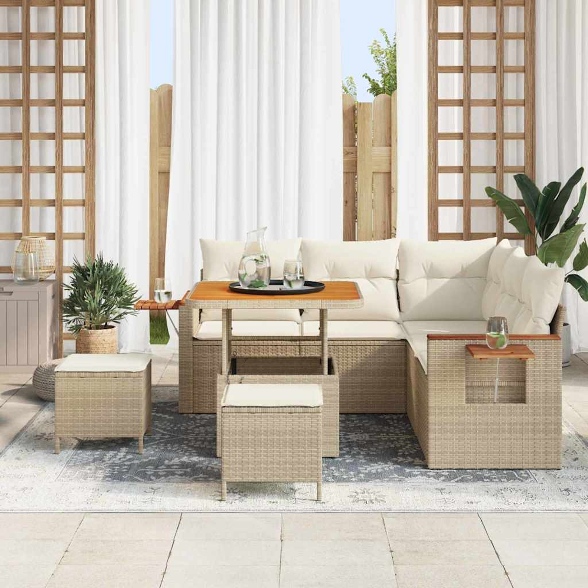 Garten-Sitzgruppe mit Kissen, Tisch und zwei Hockern. Auf dem Tisch stehen eine Kanne und Gläser. Weiße Vorhänge und Pflanzen im Hintergrund.
