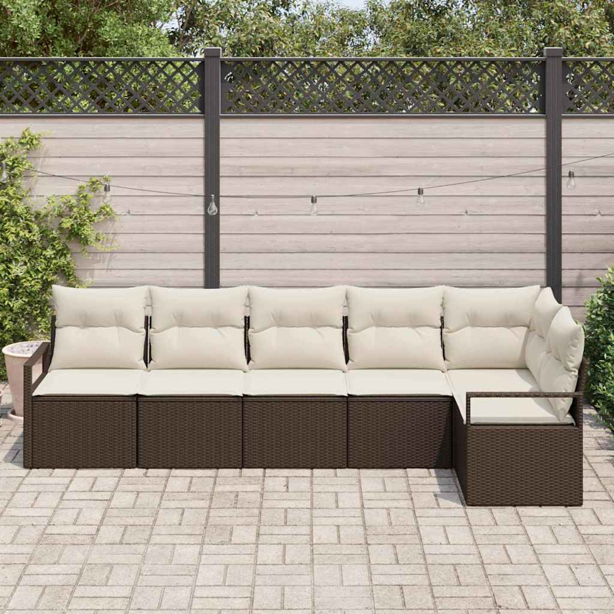 Ein Ecksofa mit cremefarbenen Kissen und braunem Korbgewebe steht auf einer Ziegelterrasse. Im Hintergrund ein Holzzaun und Grün.