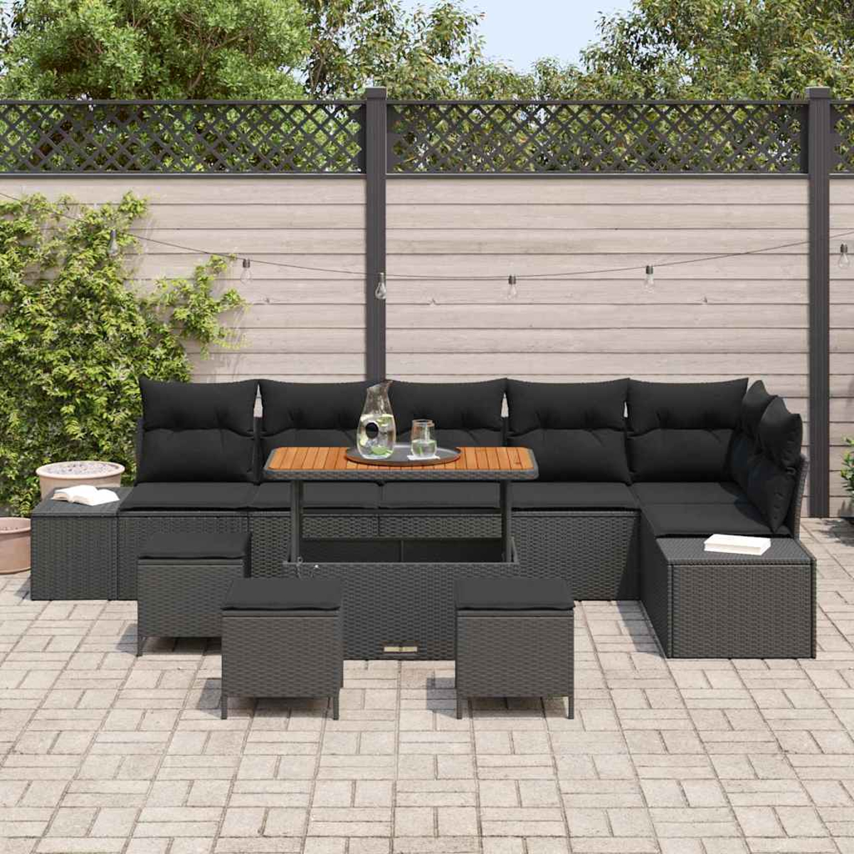 Terrassen-Set im Freien. Graues Korbmöbel mit schwarzen Kissen. Holztisch mit Glas. Blumentöpfe, graue Pflastersteine und Holzzaun.