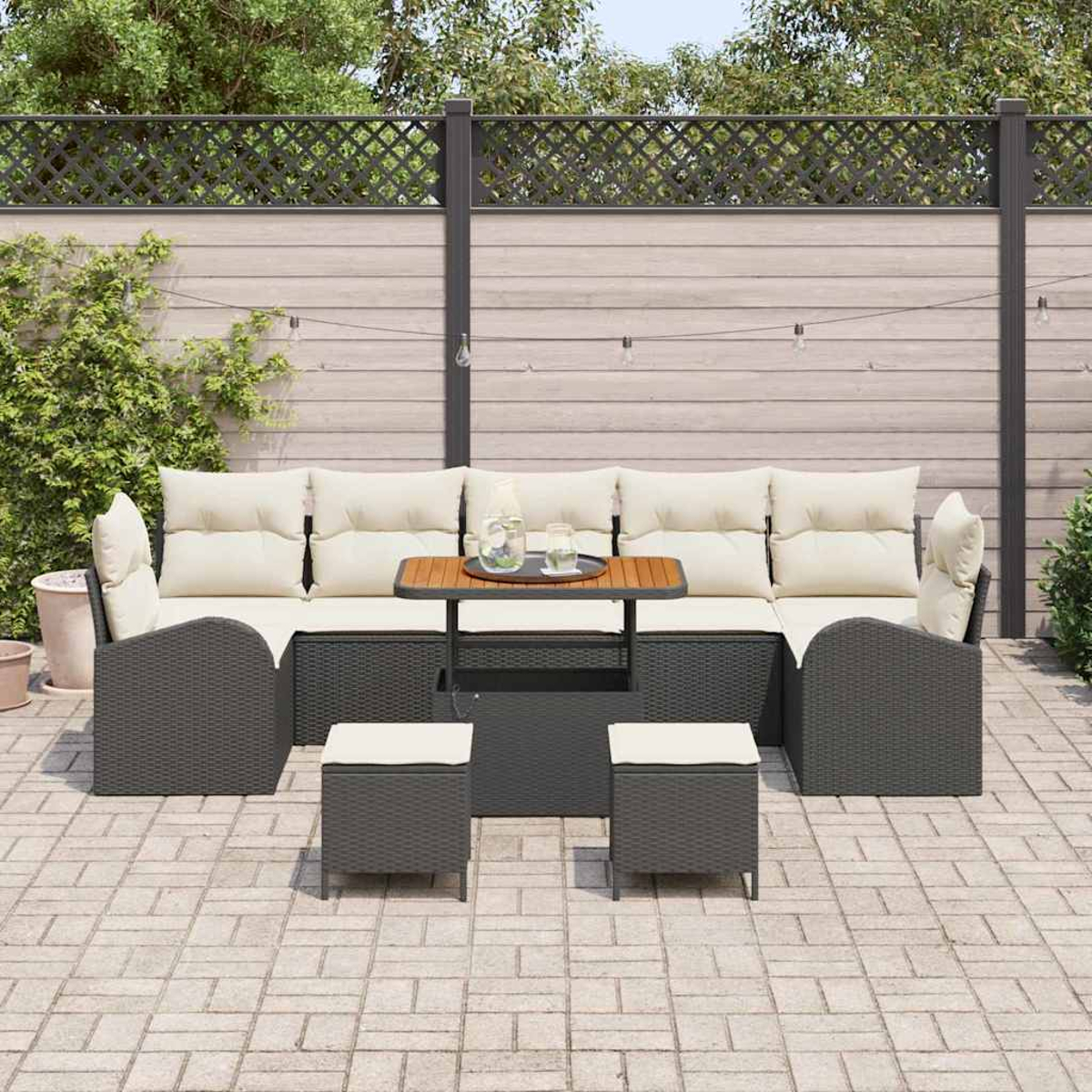 Juego de patio al aire libre con muebles de mimbre gris, cojines crema, mesa pequeña con bebidas y dos otomanas, sobre un patio de ladrillos.