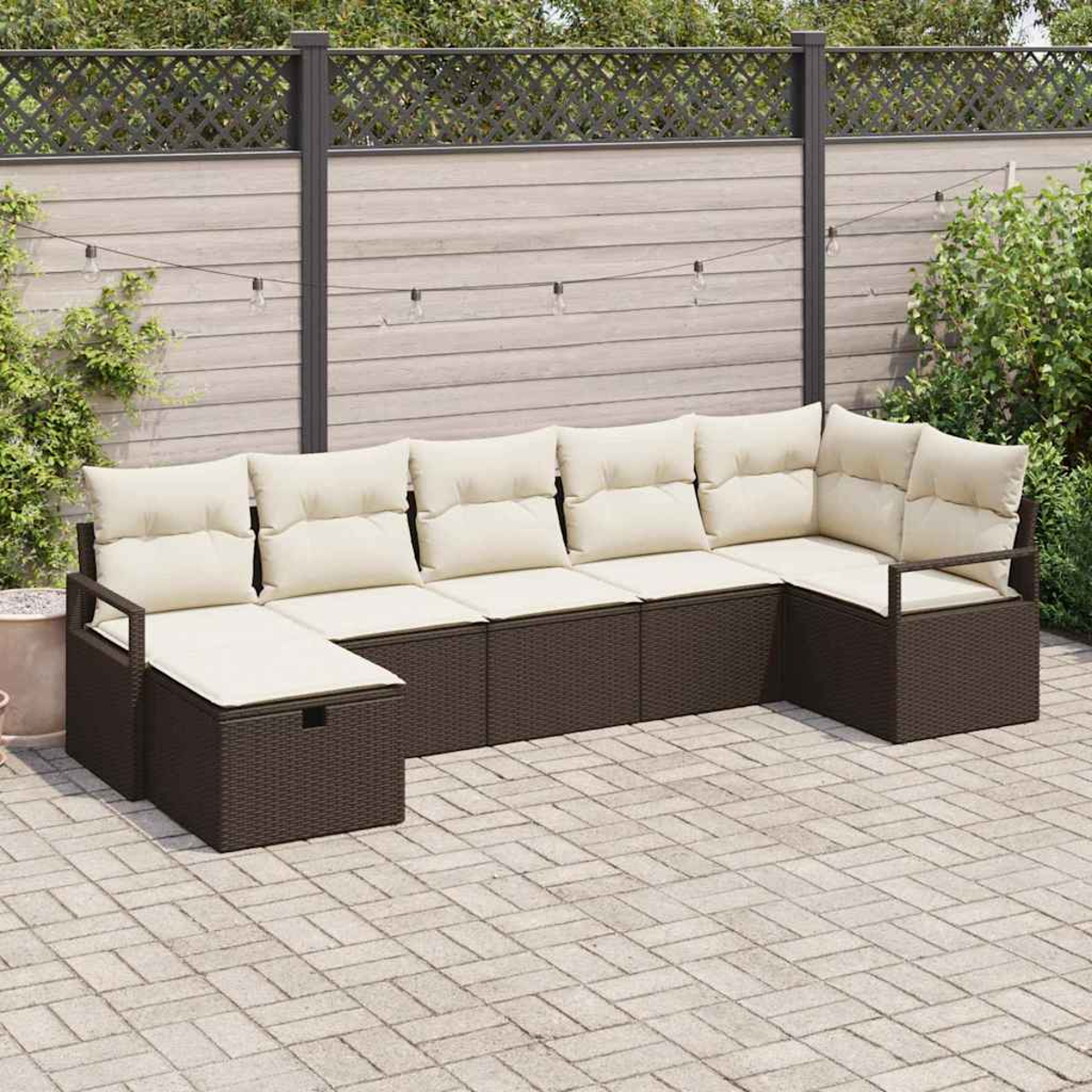 Ein braunes Outdoor-Ecksofa mit cremefarbenen Kissen steht auf einer Ziegelstein-Terrasse. Im Hintergrund sind ein Holzzaun und Lichterketten zu sehen.