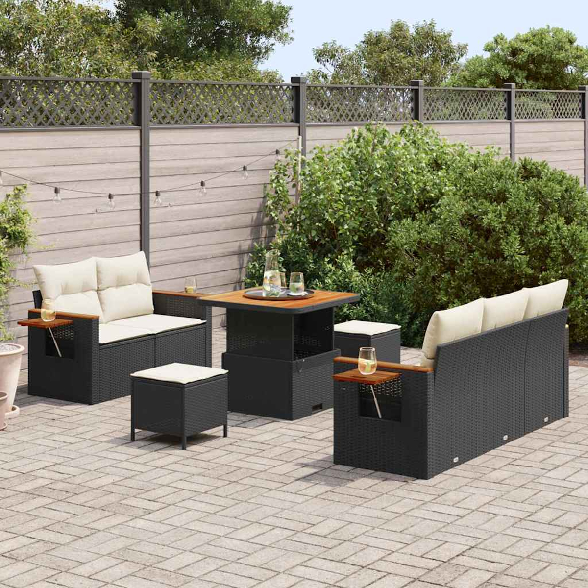 Conjunto de patio al aire libre con muebles negros tejidos, cojines beige, detalles de madera y mesa cuadrada. Patio de ladrillos, vegetación, valla.