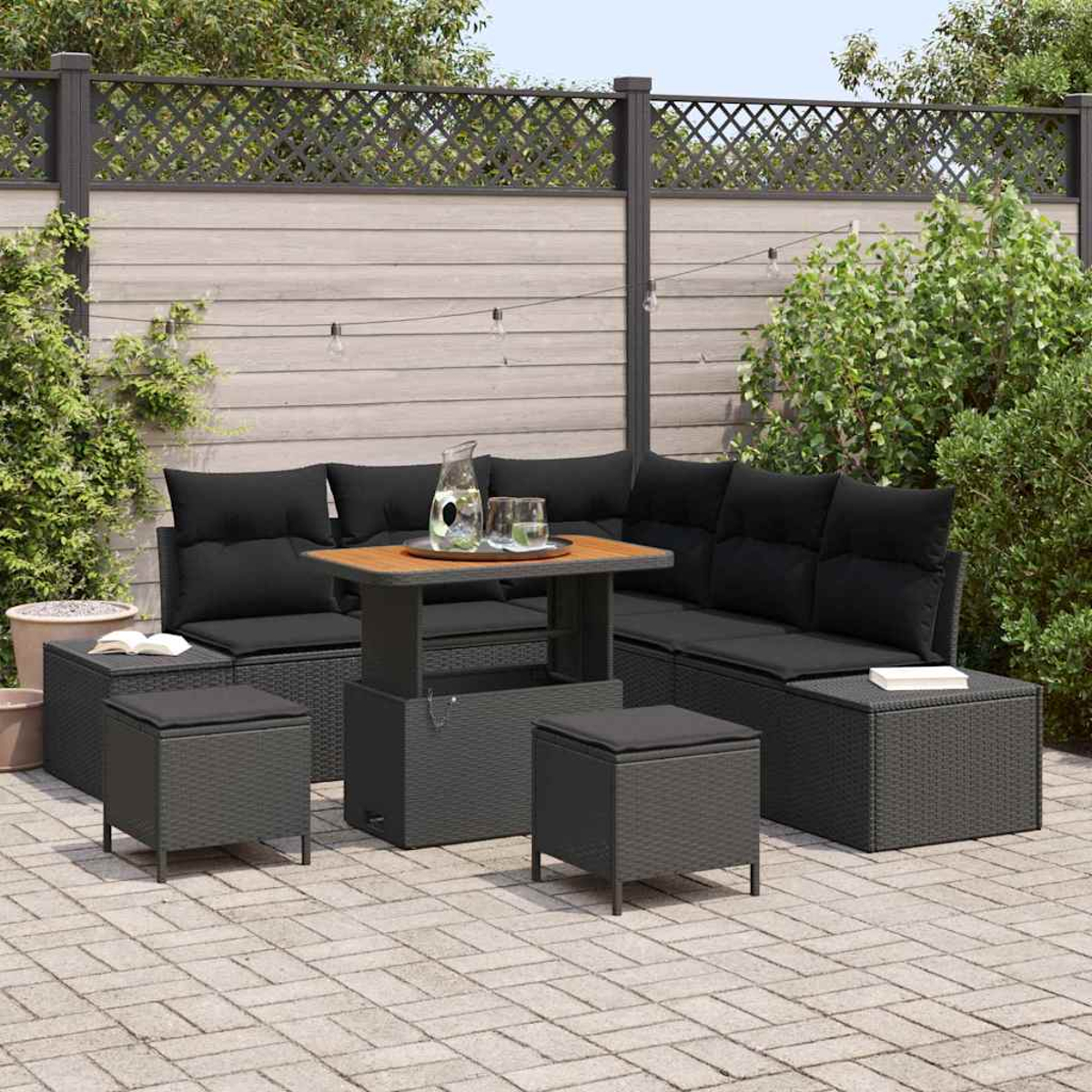 Terrassen-Set mit schwarzen Kissen, Holztisch und Hockern. Szene vor Holzzaun und Grün.