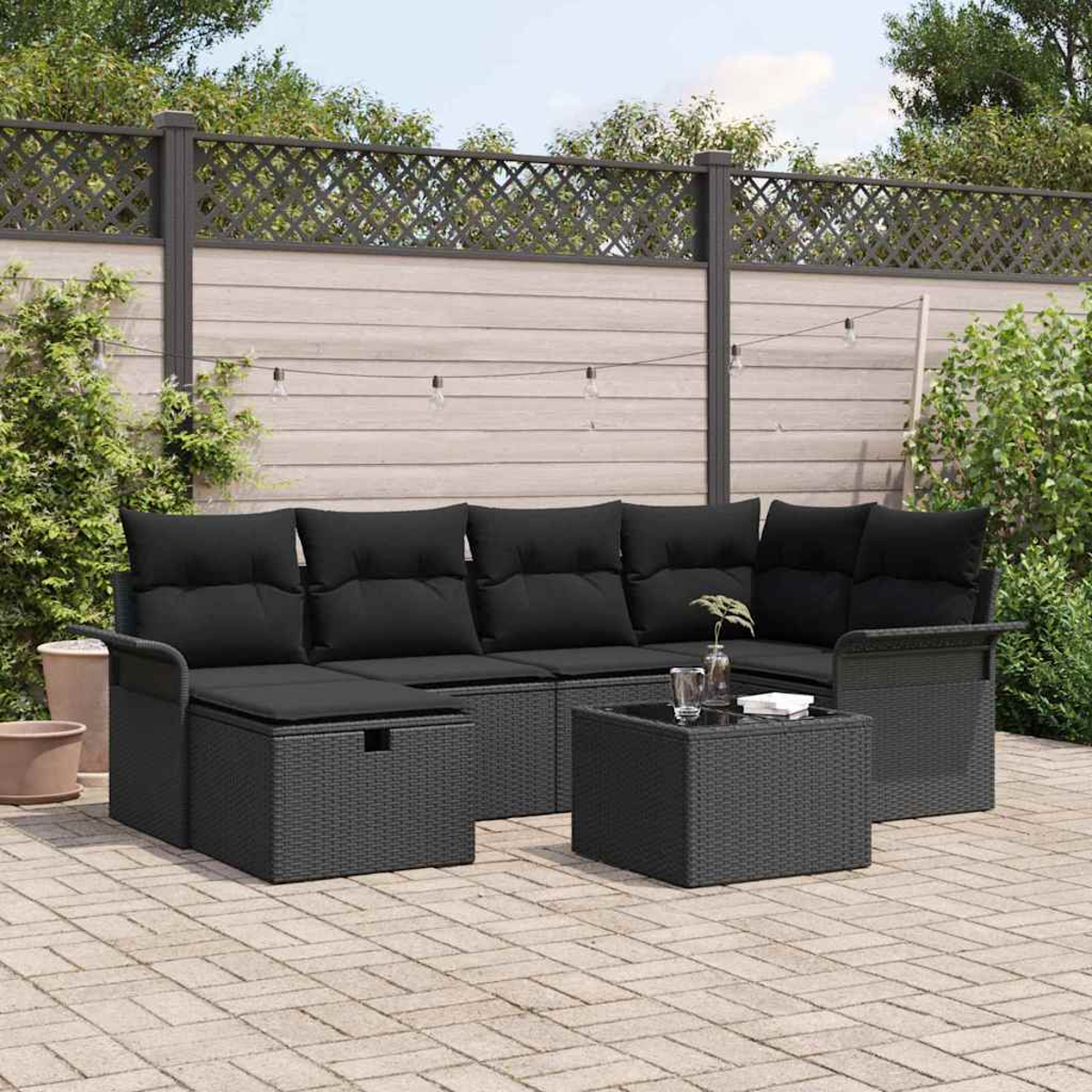 Gartenmöbel-Set: Schwarzes Rattan-Sofa mit Kissen, kleiner Tisch, Ziegelpflaster, grüne Pflanzen und Bäume im Hintergrund.