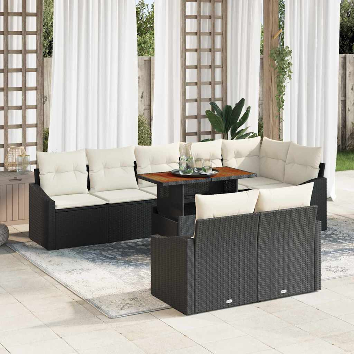 Terrassenmöbel-Set mit schwarzem Korbmöbel, weißen Kissen und einem Holztisch, vor weißen Vorhängen und einem Holzgitter.