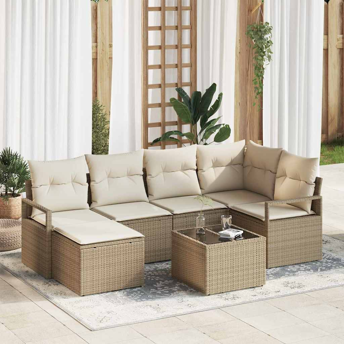 Garten-Korbsitzgruppe mit cremefarbenen Kissen, Couchtisch mit Glasplatte und Pflanzen. Im Hintergrund weiße Vorhänge und Holzgitter.