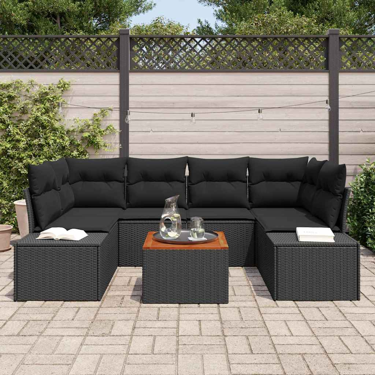 Schwarzes Outdoor-Ecksofa aus Korbgeflecht mit schwarzen Kissen, einem kleinen Tisch, Büchern und einem dekorativen Zaun im Garten.