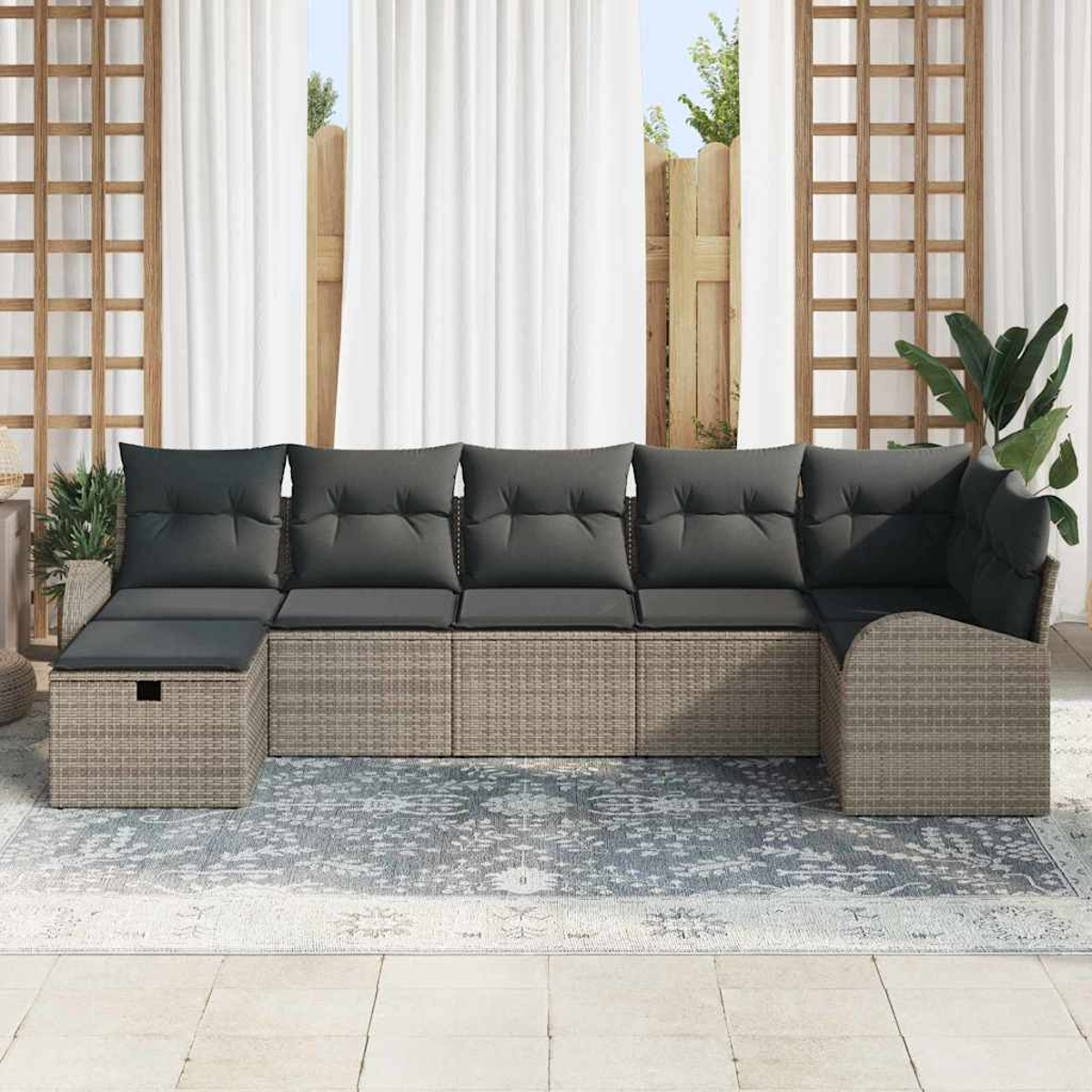 Ein graues Ecksofa mit dunklen Kissen steht auf einem gemusterten Teppich vor weißen Vorhängen und einem Holzgitter.