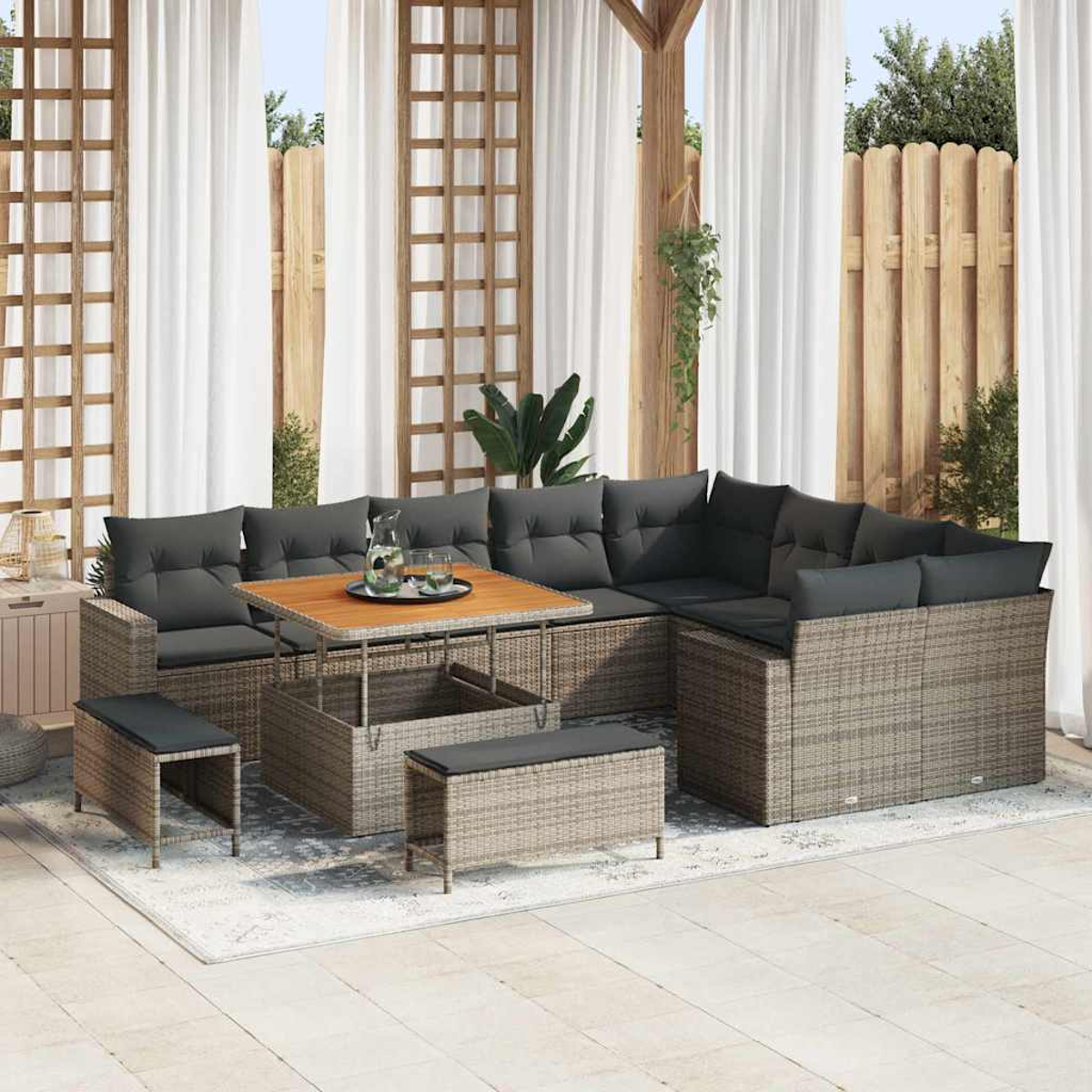 Outdoor-Rattan-Ecksofa mit grauen Kissen, Holztisch und passenden Bänken. Weiße Vorhänge und ein Holzzaun sind im Hintergrund.