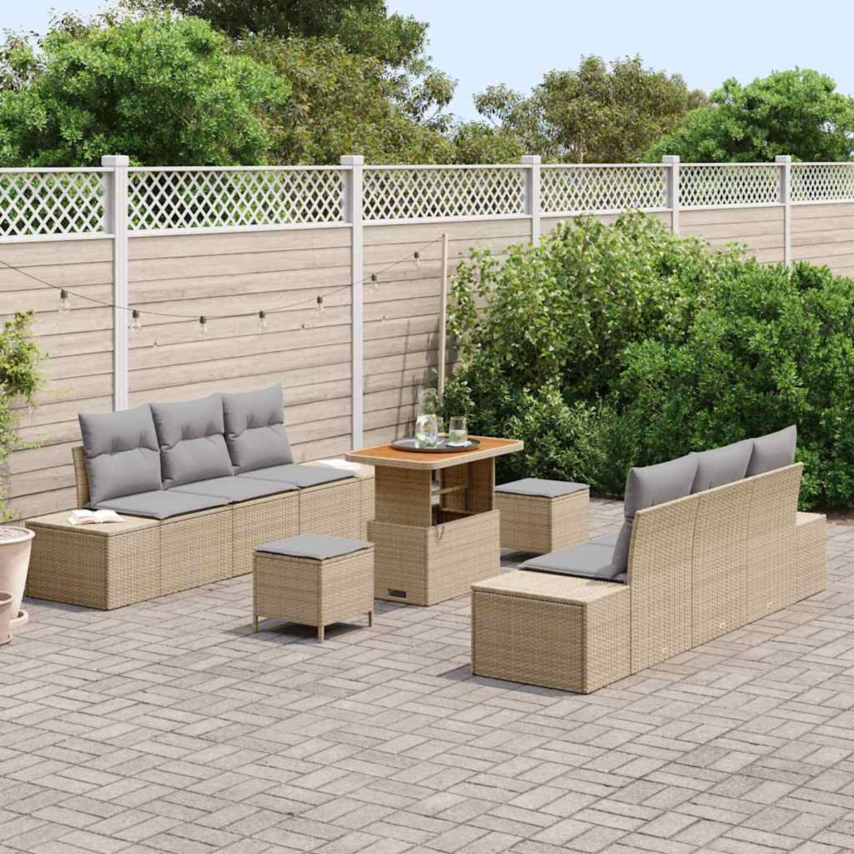 Ein Terrassenset mit beigen Korbmöbeln und grauen Kissen. Enthält ein Sofa, zwei Hocker und einen Tisch vor einem Zaun und Grün.