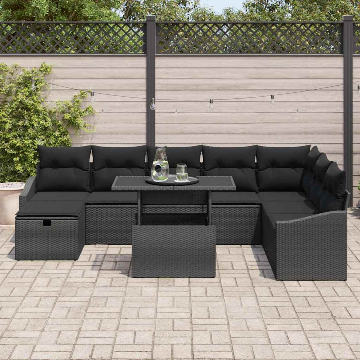 Outdoor-Ecksofa mit schwarzen Kissen und einem kleinen Tisch mit Tablett auf einer Backstein-Terrasse. Zaun und Grün im Hintergrund.