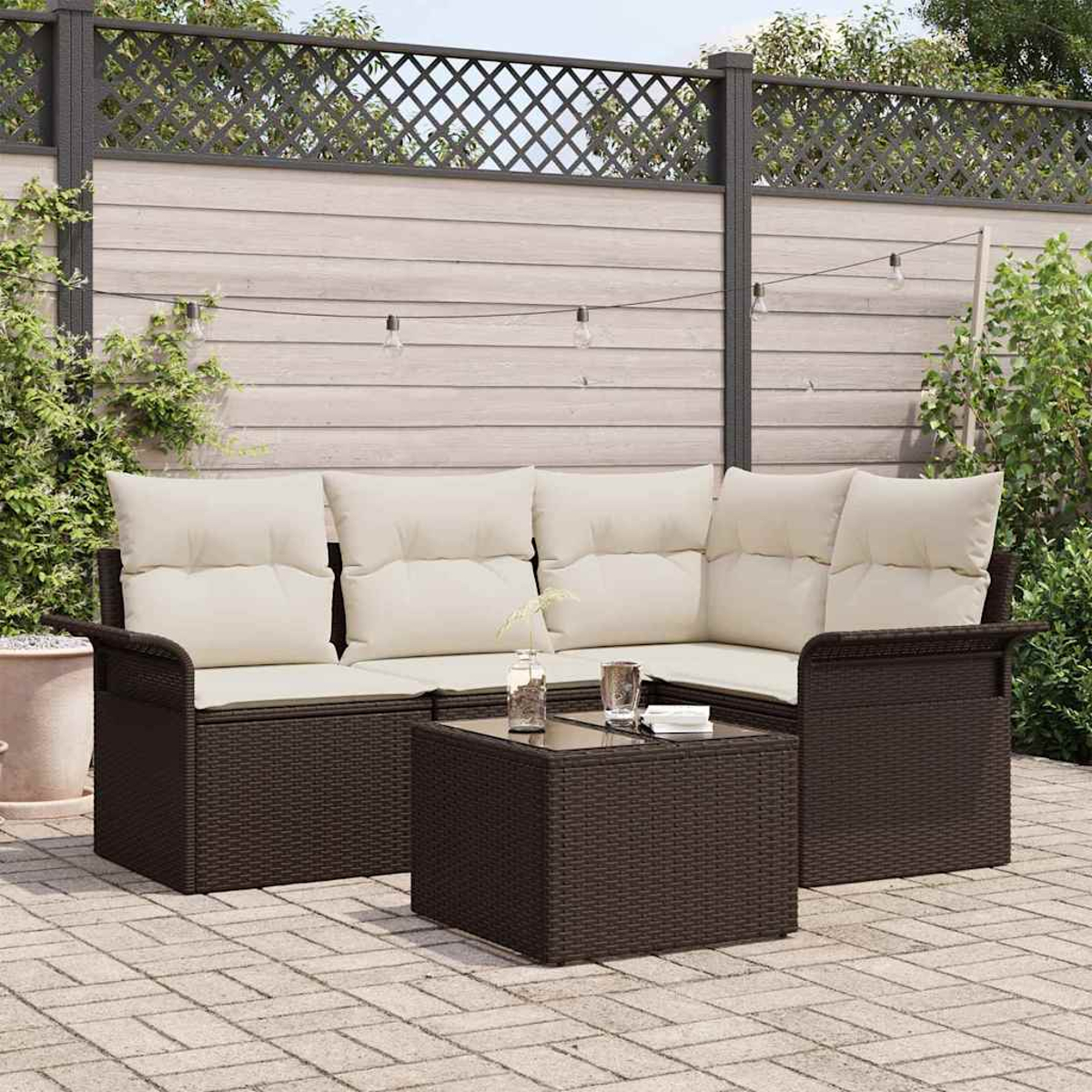 Braunes Outdoor-Ecksofa aus Korbgeflecht mit cremefarbenen Kissen und einem quadratischen Tisch. Im Hintergrund ein Holzzaun und Pflanzen.
