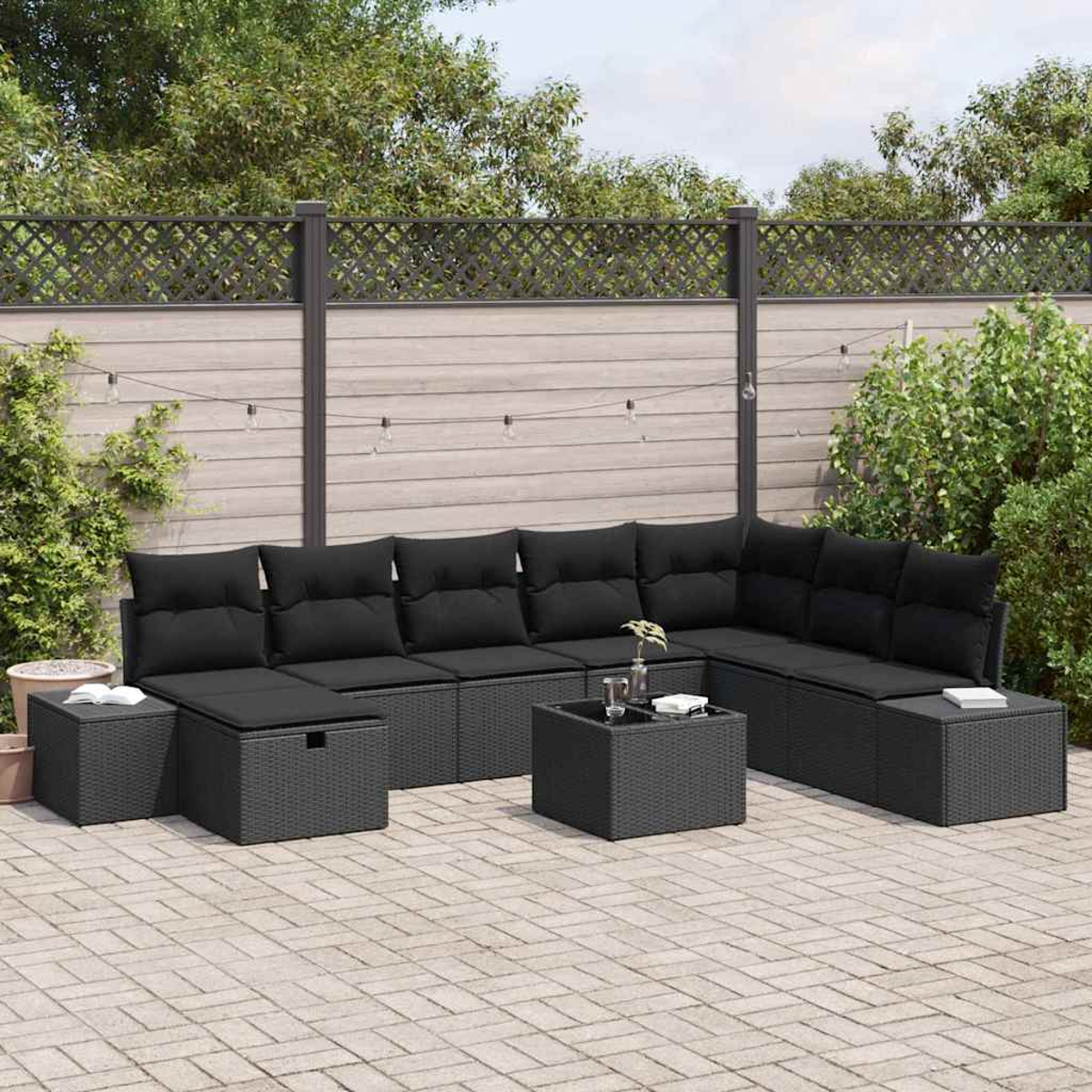 Ein schwarzes Outdoor-Geflecht-Ecksofa mit Kissen und einem kleinen Tisch auf einer Ziegelterrasse. Grüne Pflanzen und ein Holzzaun im Hintergrund.