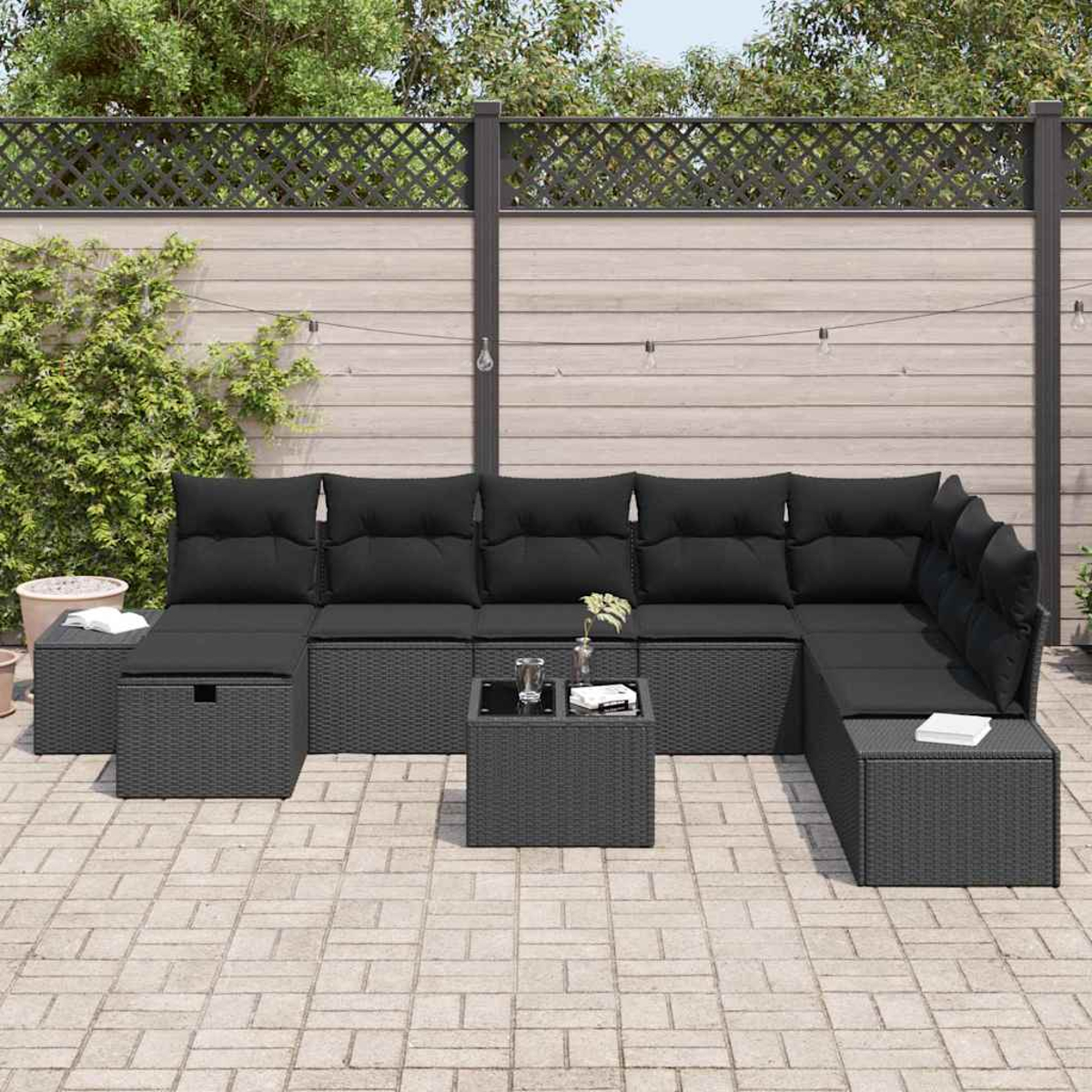 Outdoor-Terrassenset mit schwarzen Kissen auf grauem Korbgeflecht. Enthält einen Tisch mit Gläsern und einem Buch, vor einem Holzzaun.