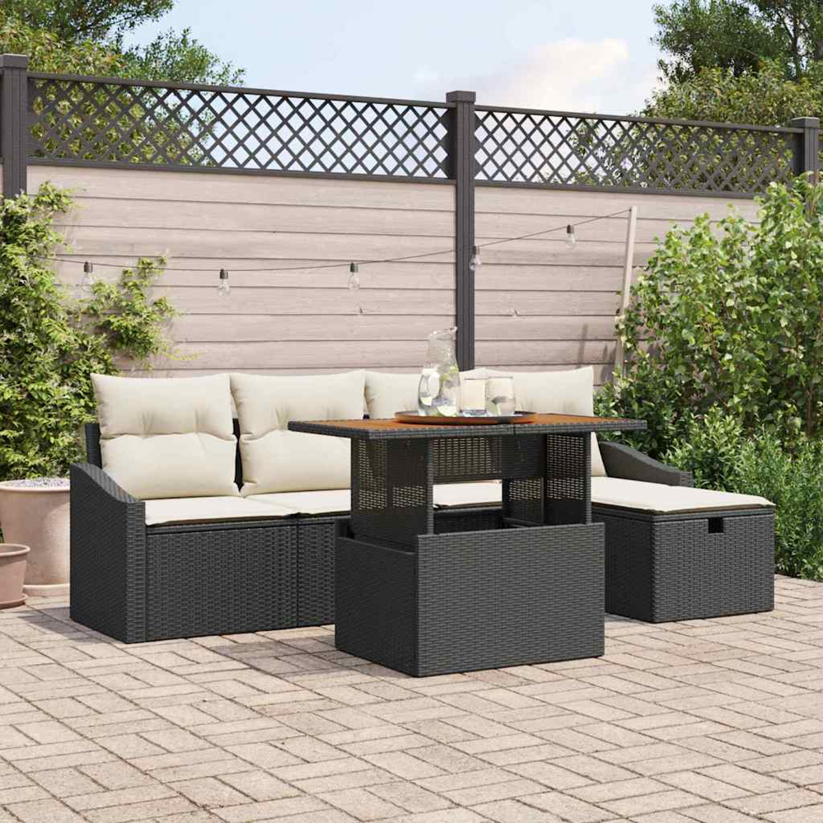 Garten-Set mit schwarzem Korbmöbel, Tisch und Stauraum-Hocker. Beige Kissen. Holzzaun mit Lichterketten im Hintergrund.